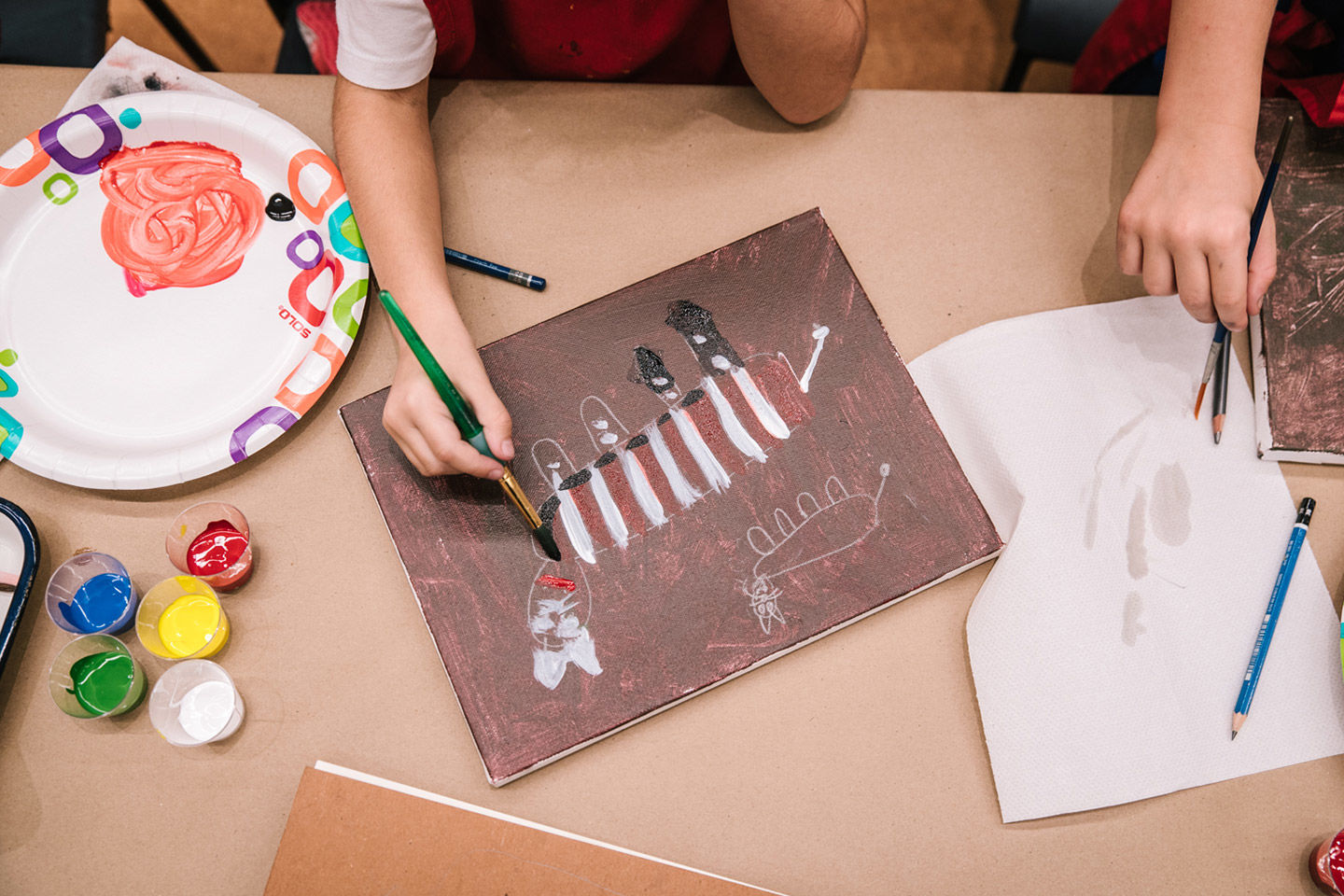 Overhead view of a small child painting a picture of an animal covered in vertical white and red stripes, set against muddy brown background; the painting is surrounded by drawing paper, a paper plate palette and small cups of blue, yellow, red, green, and white paint